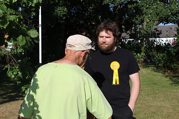 Two men facing each other in an orchard. One facing away from the camera, the other is wearing a Lib Dem rosette and facing towards the camera.
